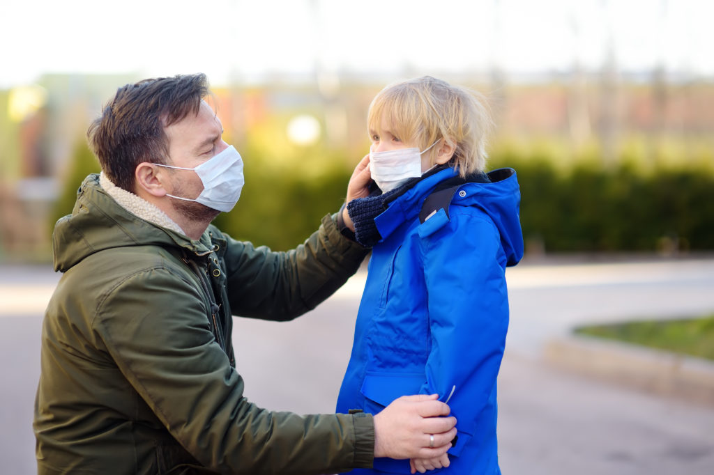 Mature man wearing a protective mask puts a face mask on a his son. Supporting caregivers like parents is important.