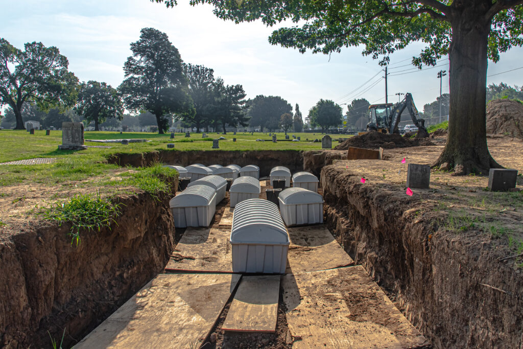 Twelve gray steel coffins rest in an earthen trench among the gravestones and mature trees of an old cemetery.