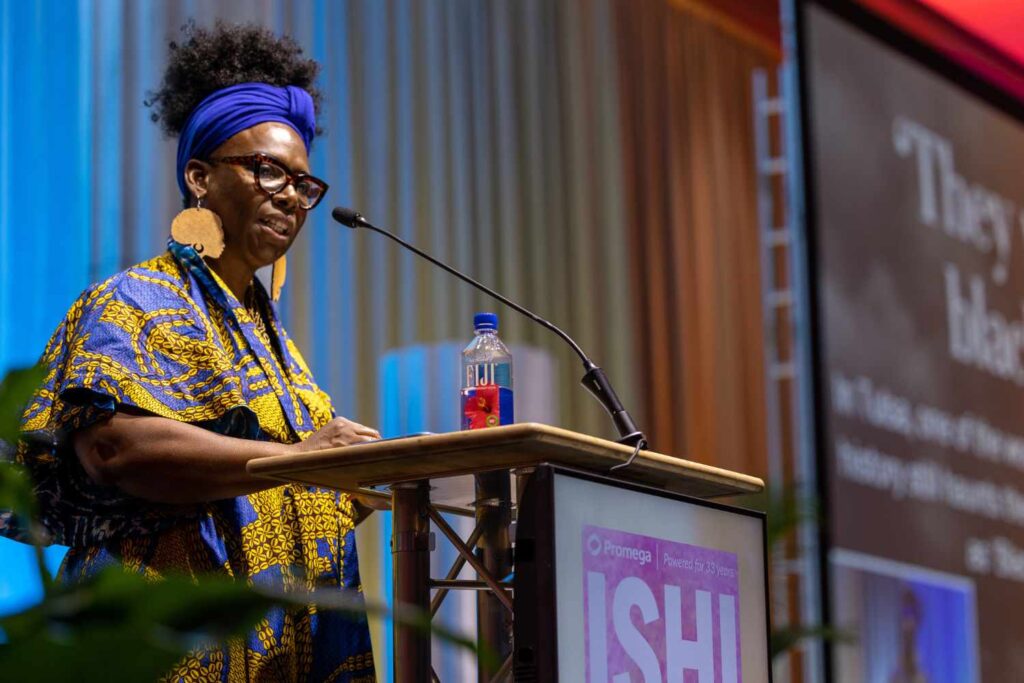 Professor DeNeen Brown, wearing a blue and yellow dress, speaks at a podium.