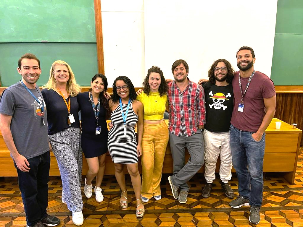 Photo of Dr. Martin Bonamino's Lab Group; eight humans, standing with arms around each other in front of a green chalk board. 