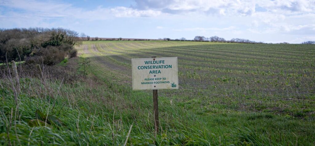 A sign reading “Wildlife Conservation Area — Please keep to marked footpaths” stands in the foreground of a grassy field, with rows of young crops and a line of trees under a partly cloudy sky.